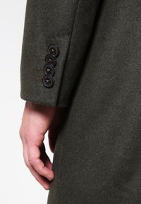 Manche de manteau en laine vert foncé avec quatre boutons sombres et des accents dorés. Texture douce, coupe ajustée, mettant en valeur un savoir-faire détaillé.