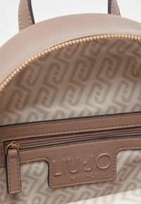 Sac à dos en cuir beige avec fermeture éclair dorée. L'intérieur comporte une doublure à motifs et une poche zippée avec logo de la marque embossé.