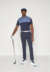 Polo com blocos de cor azul e azul-marinho, calças justas azul-marinho, cinto branco e sapatos de golfe brancos. A pessoa segura um taco de golfe e usa uma boné.