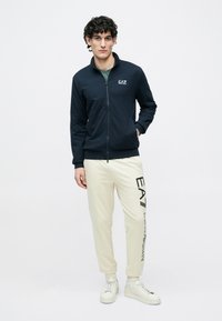 Námornícka mikina na zips s vreckami, s logom na hrudi, v kombinácii s krémovými teplákmi s vertikálnou potlačou loga, nosená s bielymi teniskami.