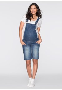 Salopette en jean, bleue, avec bretelles ajustables, poche avant et ourlets retroussés. Portée sur un T-shirt blanc avec un motif de cœurs.