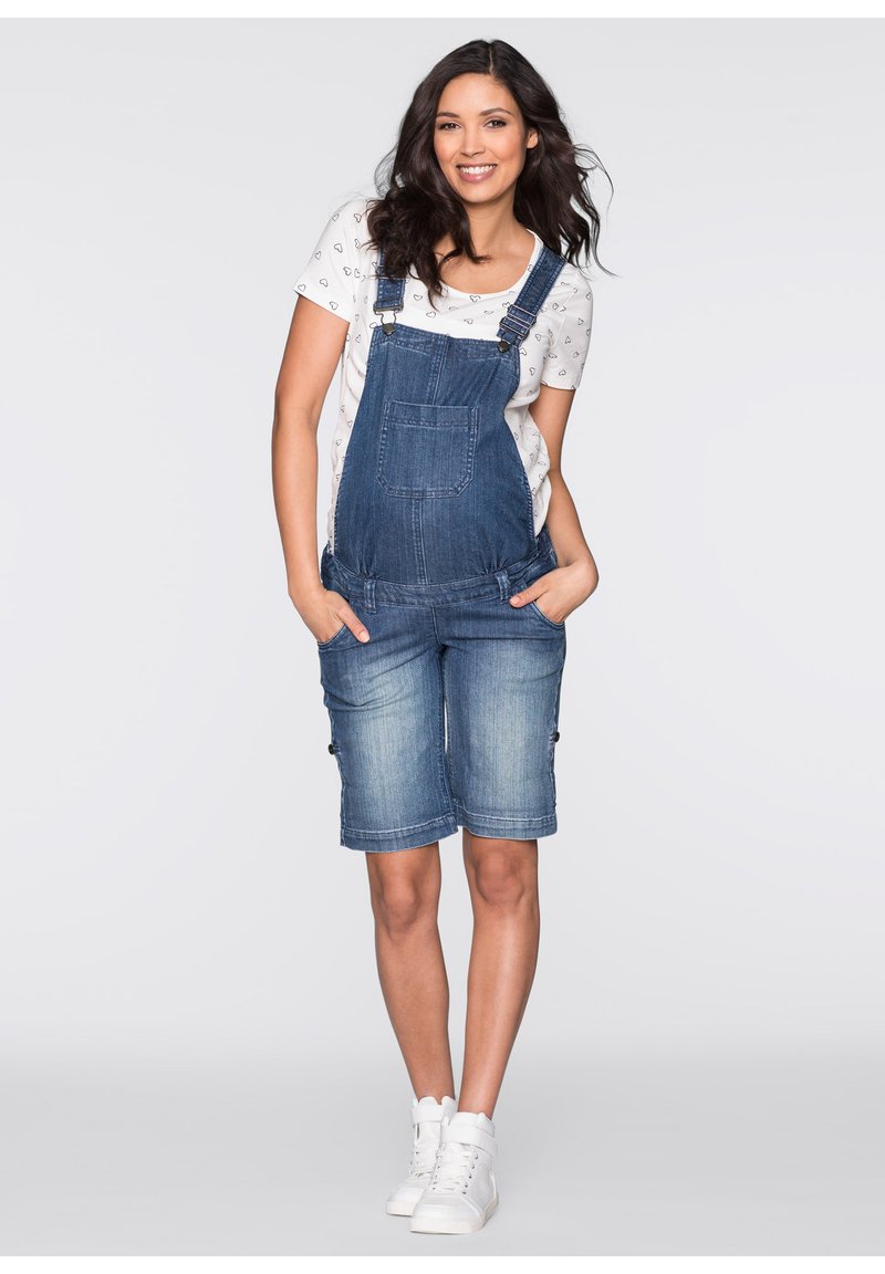 Salopette en jean, bleue, avec bretelles ajustables, poche avant et ourlets retroussés. Portée sur un T-shirt blanc avec un motif de cœurs.