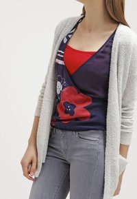 Ljusgrå, texturerad kofta över en mörk blommig topp med röda och blå detaljer, tillsammans med grå, figursydda jeans. Enkelt mönster, avslappnad passform.