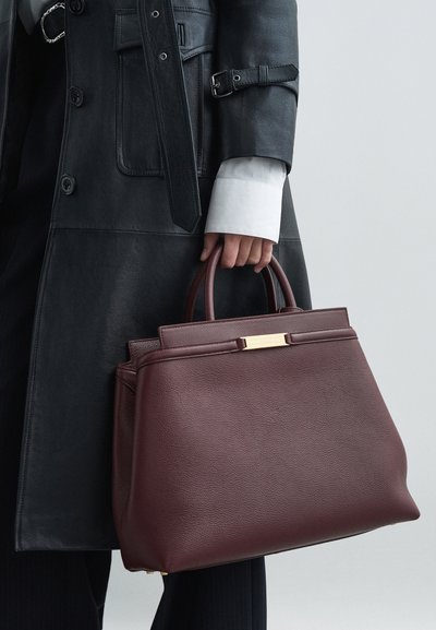 Burgundia nahast käekott struktuurse kujuga, millel on kuldne riistvara ja kaks käepidet, kantud musta nahktagi ja valge särgiga.