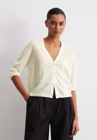 Helles gelbes, leichtes Strickjäckchen mit tiefem V-Ausschnitt, langen Ärmeln und Knopfleiste. Kombiniert mit schwarzen, weit geschnittenen Hosen.