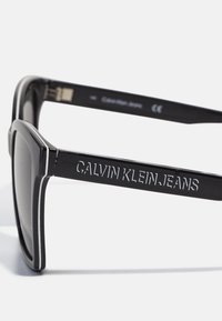 Ochelari de soare rectangulari negri cu ramă din plastic lucios, având inscripționat pe tempă "Calvin Klein Jeans". Lentile gri cu un design elegant.