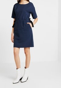 Kortärmad klänning i marinblått med texturerad yta, dragsko i midjan och svarta ribbonsaccentuerade detaljer. Matchad med vita boots i westernstil.