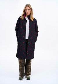 Pletený čierny kabát s červenými bodkovými akcentmi, s patentným zapínaním, postrannými vreckami a ružovou podšívkou, kombinovaný so nohavicami s leopardím vzorom.