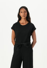 Svart ribbestrikket jumpsuit med korte ermer, rund halslinning og belte i midjen. Har en avslappet passform og sidelommer for ekstra funksjonalitet.