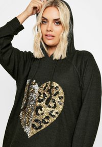 Mörkgrön huvtröja med ribbad textur och en stor paljettformad hjärt-design som har guld-, silver- och leopardskimrande detaljer.