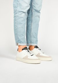 Bílé kožené a šedé semišové tenisky s černými akcenty, s designem na šněrování a silnou béžovou podrážkou. Džínové kalhoty mírně vyhrnuté na manžetách.