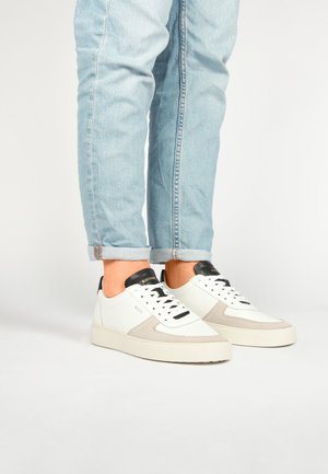 Witte leren en grijze suède sneakers met zwarte accenten, voorzien van een vetersluiting en een dikke beige zool. Spijkerbroek lichtjes opgerold bij de manchetten.