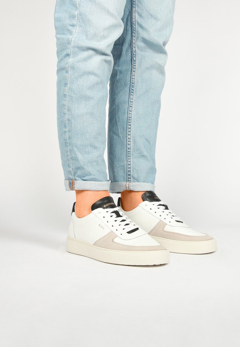 Bílé kožené a šedé semišové tenisky s černými akcenty, s designem na šněrování a silnou béžovou podrážkou. Džínové kalhoty mírně vyhrnuté na manžetách.