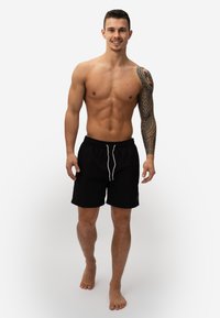 Zwarte zwembroek met een trekkoord in de taille, lichtgrijze accenten en een relaxte pasvorm; model is shirtloos met een tatoeage op zijn arm.