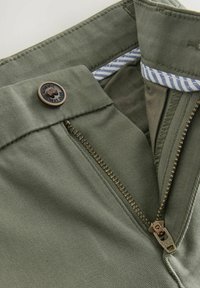 Pantaloni in cotone verde oliva con zip, dotati di chiusura con bottone in bronzo e dettaglio a righe sulla cintura interna.