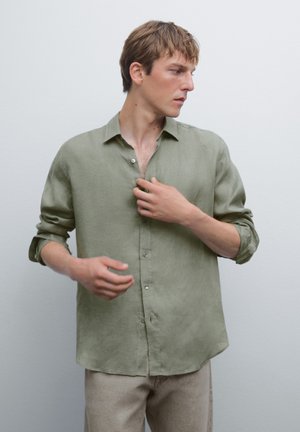 Chemise en lin vert clair avec une coupe décontractée, devant à boutons et manches retroussées. Modèle ajustant un bouton, portant un pantalon beige.