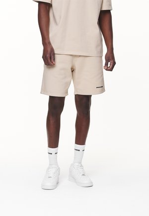 Männliches Model trägt beige Shorts und ein passendes T-Shirt, weiße Socken mit schwarzem Logo und weiße Sneaker, während es vor einem weißen Hintergrund steht.