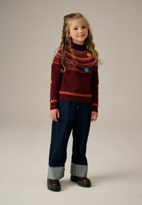 Burgundowy sweter z dzianiny z kolorowymi geometrycznymi wzorami i filcowymi aplikacjami w kształcie kwiatów, w zestawieniu z ciemnoniebieskimi dżinsami z podwiniętymi nogawkami i brązowymi butami.