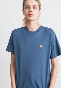 Giovane con i capelli corti biondi, che indossa una maglietta blu larga con un piccolo logo giallo Carhartt sul petto, in piedi davanti a uno sfondo bianco.