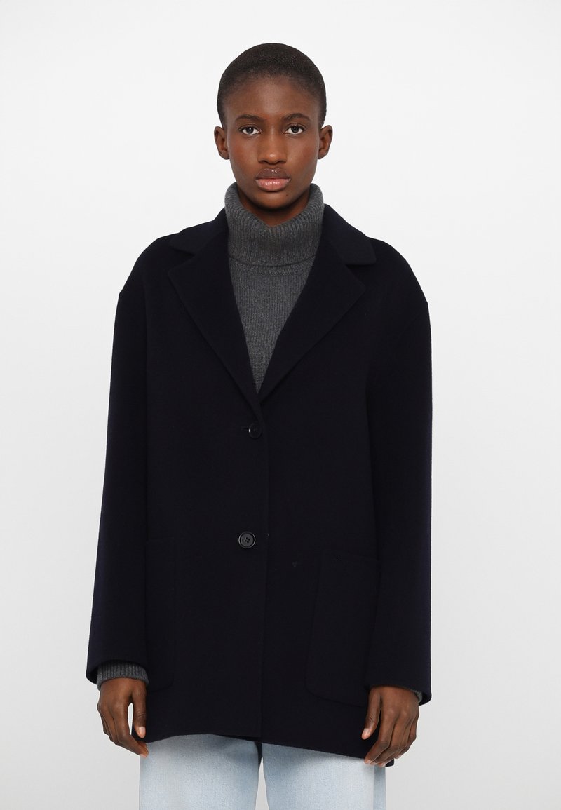 Manteau en laine bleu marine avec un col, deux poches avant et un bouton de fermeture. Porté par-dessus un col roulé gris, avec une coupe décontractée.