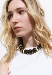 Collier en perles avec des perles rondes vertes, blanches et brunes avec des accents dorés, porté contre un top blanc uni et des cheveux légers et ondulés.
