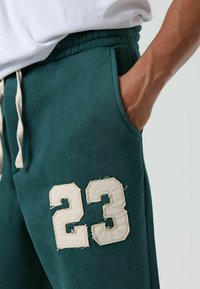 Groene joggingbroek van zachte stof, met een verweerde crème nummer "23" patch en een taille met trekkoord in lichtbeige kleur.