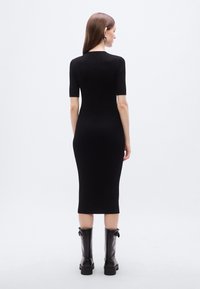 Μαύρο μίντι φόρεμα με ριμπ ύφανση, κοντά μανίκια και εφαρμοστή σιλουέτα. Το μοντέλο φοράει μαύρες μπότες μέχρι το γόνατο με χοντρό τακούνι.