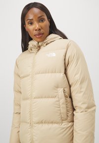 Giacca piumino beige con chiusura a zip, cappuccio e logo sul petto. Presenta una superficie trapuntata e tasche laterali. Finitura liscia.