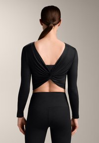 Ženska s čopom, oblečena v črno kratko majico z zasukom na hrbtu in visokim pasom legic, stoji pred enakomernim nevtralnim ozadjem.