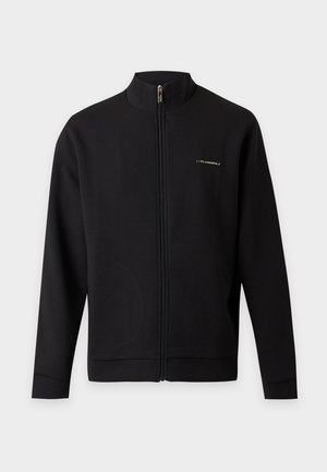 Homme portant une veste noire zippée, un pantalon beige et des baskets noires, debout les mains dans les poches devant un fond clair uni.
