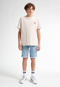 Béžové bavlněné tričko s malým růžovým motivem na hrudi, v kombinaci s ohrnutými světle modrými džínovými šortkami a bílými teniskami s černými detaily.