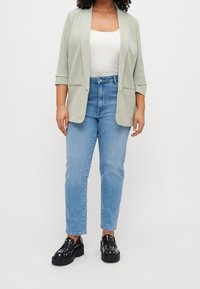 Lichtgroene blazer met driekwartmouwen, witte tanktop, lichtblauwe hoge taille jeans en zwarte platform loafers met studs.