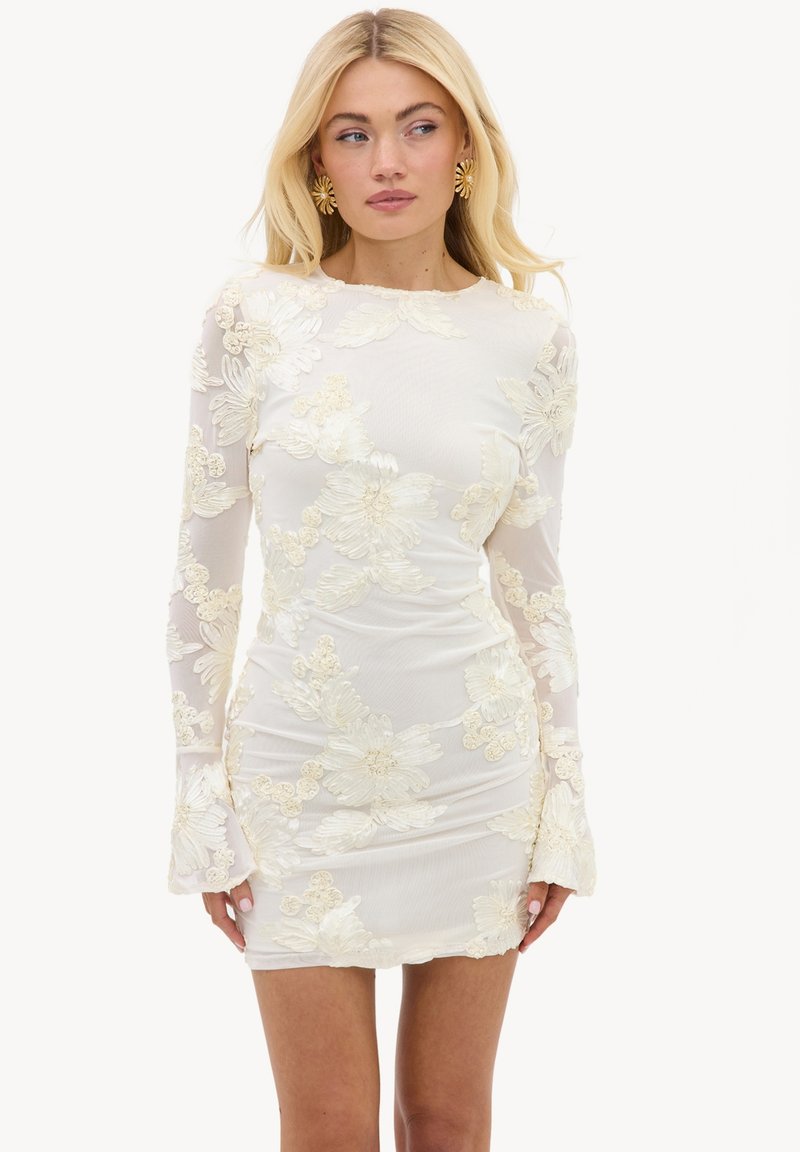 Femme blonde portant une mini-robe blanche transparente et ajustée avec une grande broderie florale et des manches longues, associée à des boucles d’oreilles dorées en forme de fleur.