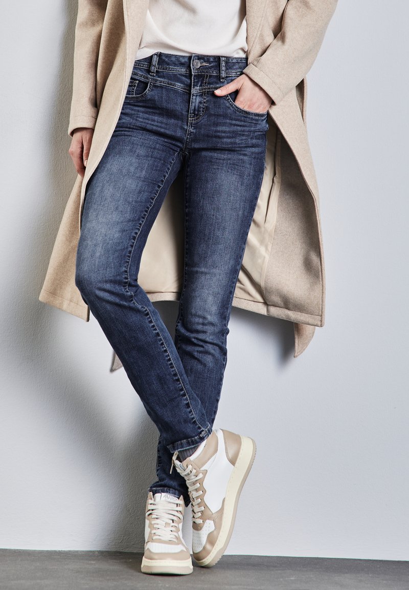 Jean bleu foncé à coupe ajustée, porté avec un manteau beige et des baskets blanches avec des accents beiges et des panneaux texturés.