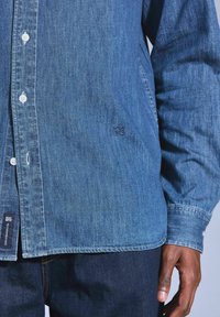 Camicia in denim di colore blu scuro con chiusura frontale a bottoni, bottoni bianchi e logo ricamato sul petto. Presenta maniche lunghe e orlo curvato.