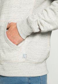 Sweatshirt gris clair en tissu texturé, avec une poche frontale, des poignets côtelés et une petite étiquette bleue avec du texte sur le côté.