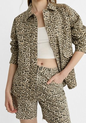 Vrouw draagt een beige en zwarte denimjas met luipaardprint en bijpassende korte broek, gecombineerd met een wit cropped tanktop.