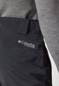 Pantalons de neige imperméables noirs avec une texture lisse, arborant un logo Columbia Titanium sur le côté et une taille sécurisée.
