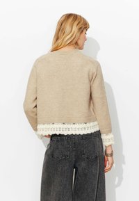 Pull beige en tricot avec un ourlet et des manches ornés d'une bordure en dentelle crème en forme de feston. La personne le porte avec un jean gris foncé à taille haute et jambes larges.