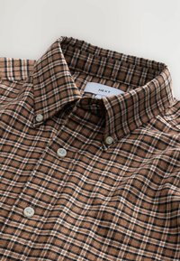 Camisa de franela a cuadros marrón y negro con cuello abotonado, que presenta botones marrones claros y una etiqueta con la inscripción "NEXT". Tejido texturizado.