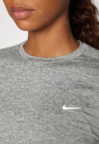 Grå, tekstureret stof crew neck skjorte med et hvidt Nike swoosh-logo på venstre brystområde. Nærbillede af halsudskæringen og den øverste del af torsoen.
