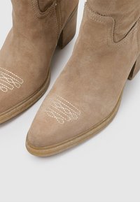 Botas de ante beige hasta el tobillo con puntera afilada y tacón cuadrado, que cuentan con un delicado bordado blanco en la zona de los dedos.