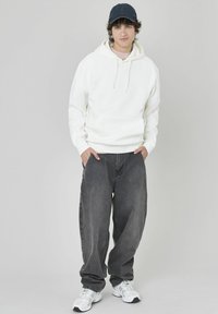 Sweat à capuche blanc, jean baggy gris et baskets blanches. Le mannequin se tient les mains dans les poches, portant une casquette de baseball bleu marine.