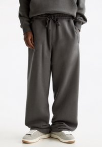 Pantalones de chándal grises con cinturilla elástica y cordón; cuentan con bolsillos laterales y un corte relajado. Combinados con zapatillas de deporte gris claro.