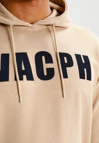 Felpa beige con lettering nero ben visibile "MACPH." Presenta un cordino con estremità argentate e un tessuto morbido e strutturato.