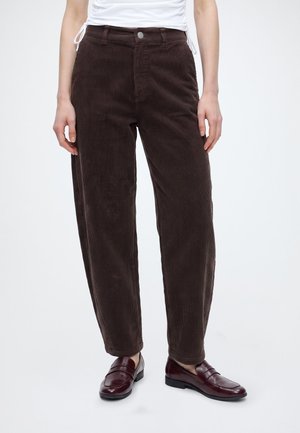 Personne portant un pantalon en velours côtelé marron foncé et des mocassins en cuir rouge foncé, debout sur un fond clair uni.