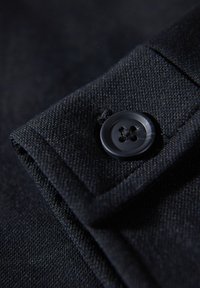 Tela negra con una superficie texturizada, que presenta un solo botón negro cosido en un puño. El botón tiene cuatro orificios y es circular.