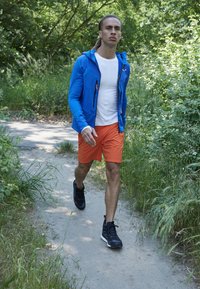 Sportslig mand i en blå lynlås hættetrøje, hvid skjorte og orange shorts, der går på en sti iført sorte træningssko, omgivet af grønne omgivelser.