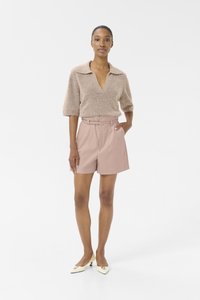 Beige gebreide trui met een V-hals en korte mouwen, gecombineerd met lichtroze shorts met een gecinste taille en een getailleerd silhouet. Witte hakken.