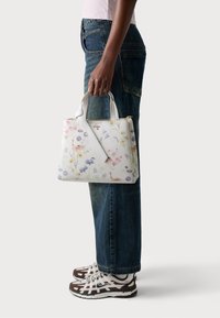 Sac à main à imprimé floral avec une surface texturée blanche, deux anses supérieures et une bandoulière amovible, associé à un jean en denim et des baskets.